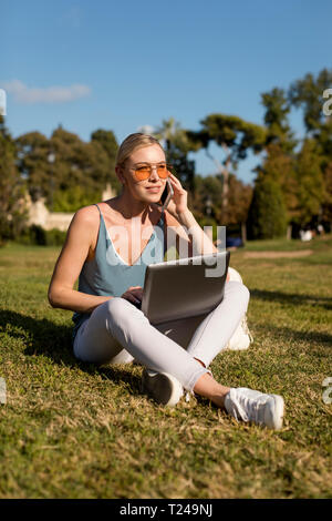 Young woman sitting in park using cell phone and laptop Stock Photo
