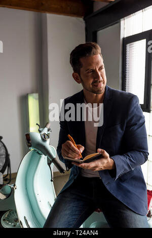 Smiling man with motor scooter in a loft writing into notebook Stock Photo
