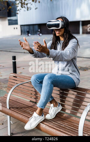Young woman sitting on a bench, using VR goggles, reaching out with her hands Stock Photo