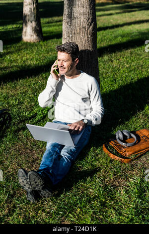 casual businessman sitting on grass in a park, using laptop, talking on the phone Stock Photo
