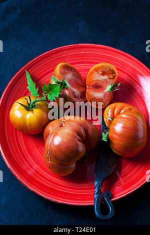 Whole and sliced beef tomatoes 'Chocolate Stripes' on red plate Stock Photo