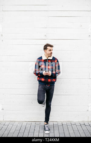 Young man with red and blue jacket using phone in a white wall Stock Photo