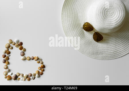 Romantic summer decoration. Heart symbol from seashells and sun hat decorated with aviator sunglasses on white background. Stock Photo