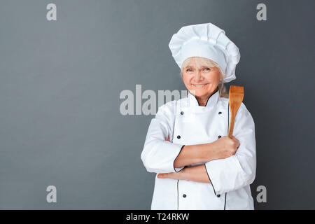 Senior female chef studio standing isolated on gray wall holding wooden spatula crossed arms looking camera smiling happy Stock Photo