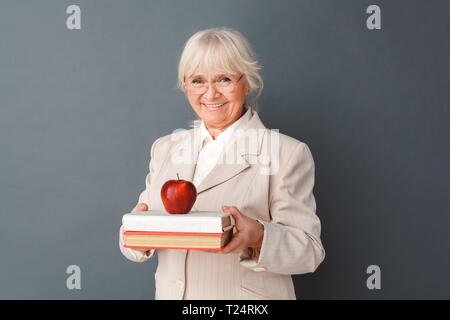 Senior woman wearing fromal suit and eyeglasses studio standing isolated on gray wall carrying books and red apple looking camera smiling positive clo Stock Photo