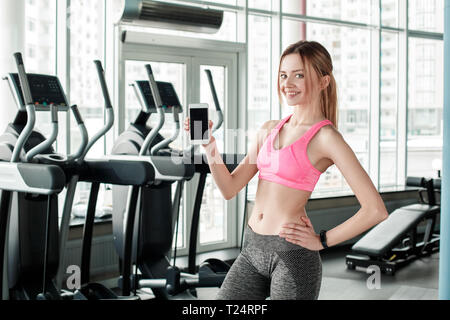 Young woman in gym healthy lifestyle standing holding smartphone showing screen looking camera smiling cheerful posing confident Stock Photo