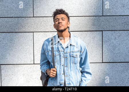 Young mulatto man carrying leather backpack standing isolated on concrete wall looking camera smiling confident Stock Photo