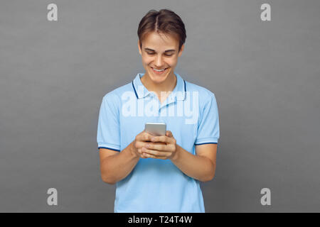Young man standing isolated on grey wall using application on smartphone smiling cheerful Stock Photo