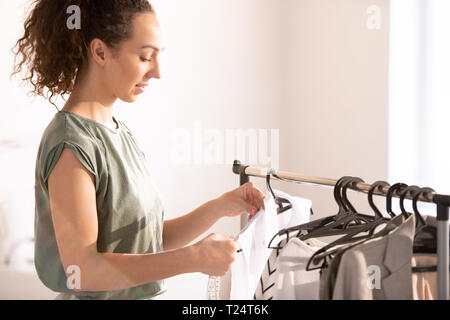 Young seamstress measuring shoulder length of new white blouse on hanger while working in her studio Stock Photo