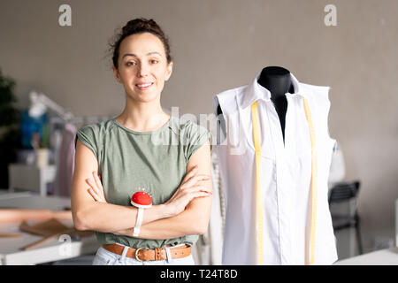 Young brunette female with her arms crossed on chest standing by dummy in front of camera in fashion studio Stock Photo