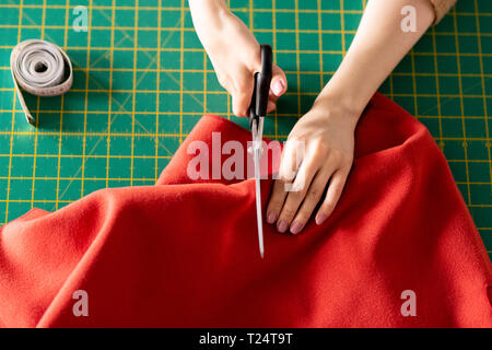 Human hands holding scissors and red textile while cutting it on green board during work in tailoring shop Stock Photo