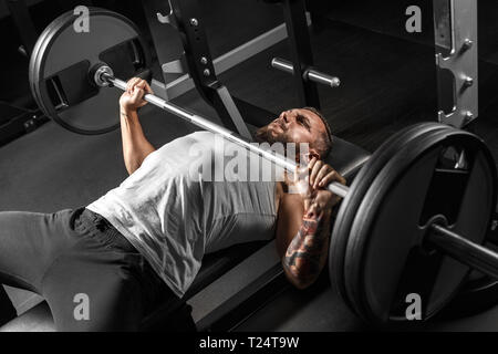 Bearded man bodybuilder exercising at gym lying doing bench press holding barbell red face making effort side view Stock Photo