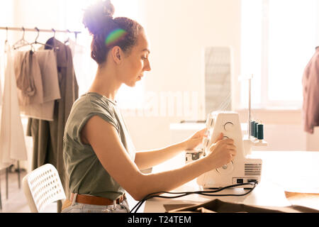 Pretty young seamstress sitting by sewing machine on table while doing her work in studio or tailoring shop Stock Photo