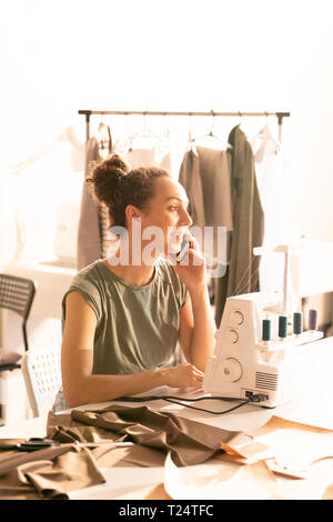 Young woman talking on the phone to client while working over new order by sewing machine in tailoring shop Stock Photo