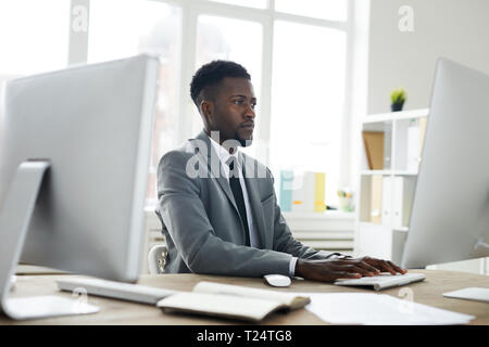 Young elegant man in formalwear looking at screen of one of computers while sitting by desk during work Stock Photo