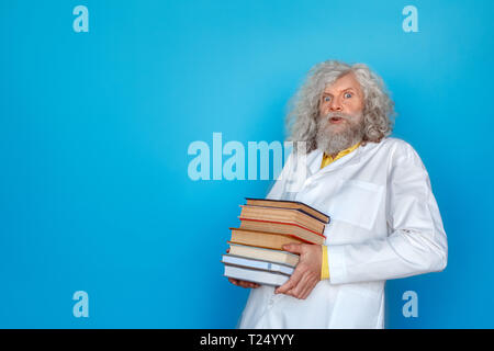 Old long-haired man wearing doctor's gown studio isolated on blue wall standing holding pile of books leaning back looking camera shocked Stock Photo