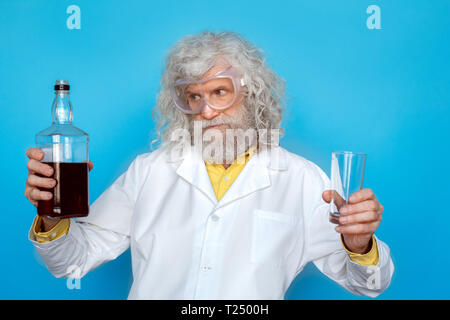 Old long-haired man wearing doctor's gown and goggles studio isolated on blue wall standing holding glass looking at bottle of spirit thoughtful Stock Photo