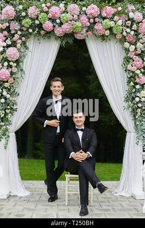 Pair of the handsome young men in white shirts and in the stylish black suits with bow tie, smiling and posing under the pink floral arch. Stock Photo
