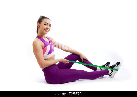 A dark-haired woman coach in a sporty purple  short top and gym makes and stretch  legs with sport fitness rubber bands, doing exercise on a  white is Stock Photo