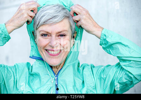 Portrait of smiling sporty mature woman putting on hood of her jacket Stock Photo