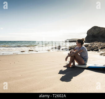 Young woman sitting on her surfboard on the beach, looking at the sea Stock Photo