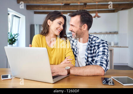 Couple sitting at dining table, using laptop, smiling Stock Photo