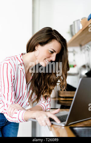 Smiling woman using laptop in kitchen at home Stock Photo