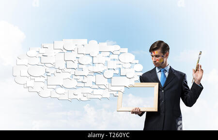 Portrait of young and successful businessman in black suit holding paintbrush in hands an looking at picture frame while standing against blue cloudy  Stock Photo
