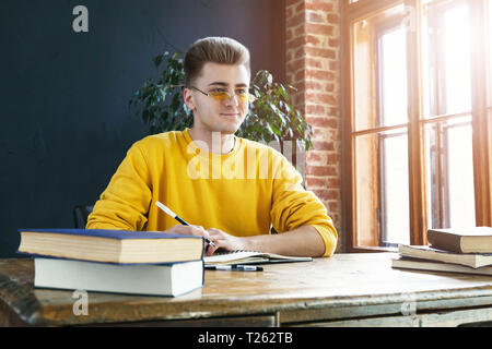Young serious blonde man wearing glasses and yellow pullover studying or working in the class Stock Photo