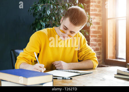 Focused blonde student in glasses making notes writing down information from book preparing for test or exam Stock Photo
