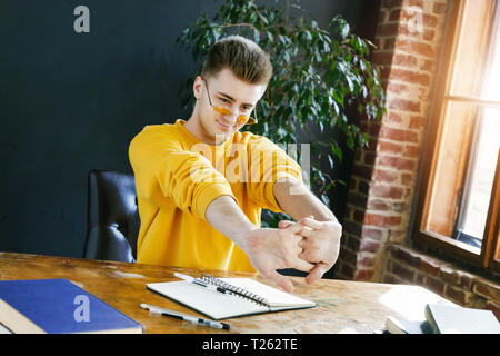 Blonde student in glasses wearing yellow pullover stretching his hands while preparing for test or exam Stock Photo