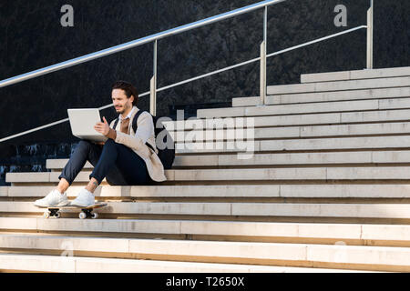 Man with backpack and skateboard sitting on stairs using laptop Stock Photo