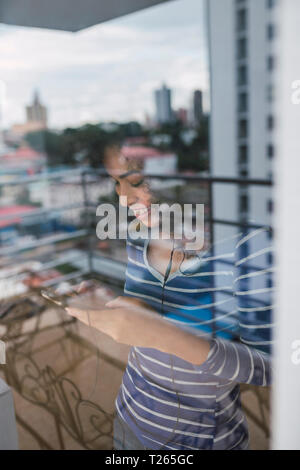 Smiling young woman behind windowpane using smartphone and earphones Stock Photo