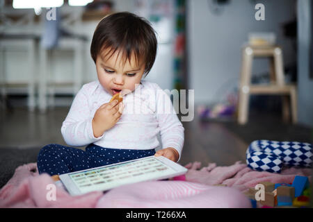 Baby girl sitting on the floor at home eating a cookie and using tablet Stock Photo
