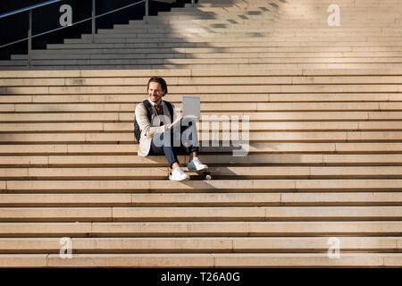 Laughing man with backpack and skateboard sitting on stairs using laptop Stock Photo
