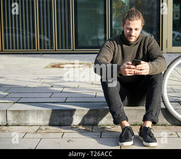 Portrait of bearded young man sitting on curb next to bicycle using cell phone Stock Photo
