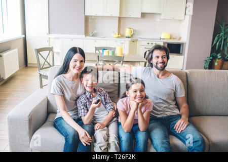 Nice and positive people from one family are sitting together on sofa. Boy leans to mother. Girl holds her hands together under chin. Guy sits in rela Stock Photo
