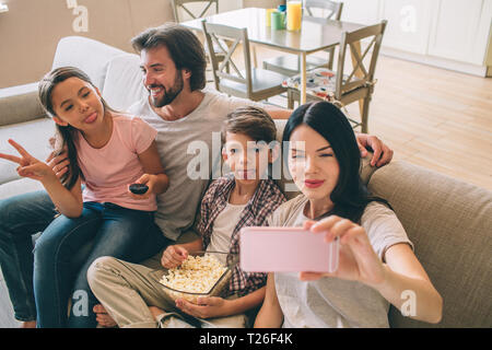 A picture of woman holds phone in hands and taking selfie of her family. Children are looking and posing. Dad smiles and looks at his daughter Stock Photo