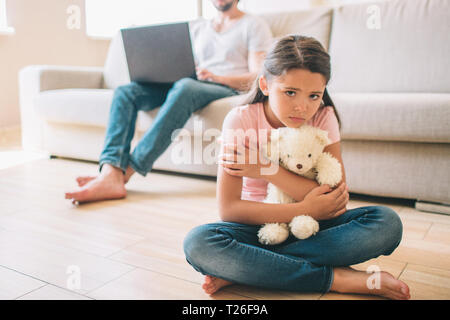 Small girl is sitting on floor. She is hugging her white bear toy. Girl looks to the right. She feels lonely. Her dad sits on sofa and work witth lapt Stock Photo