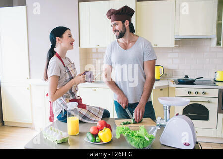 Happy man and woman stand in kitchen and look at each other. Guy cuts cucumber with knife. Young woman holds cup of tea in hands. They are smiling Stock Photo