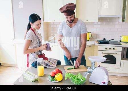 Man and woman are standing in kitchen. Guy is cutting cucumber. Girl looks at it. She holds cup of tea. They are happy Stock Photo