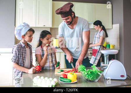 Positive dad cuts tomato with knife. Girl holds cucumber and eats it. Boy has tomato in hands and looks at what dad is doing. Mother stands behind the Stock Photo