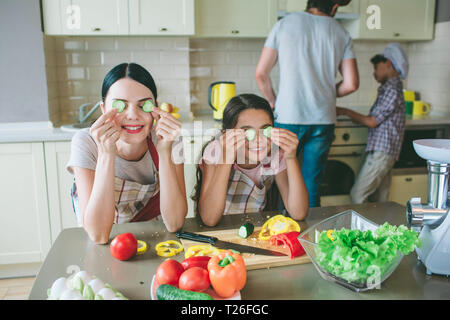 Funny girls are playing with food. They holds round pieces of cucumber where etes are and smile. Girls have a break. Biys are working at stove. They c Stock Photo