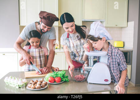 Man helps her daughter to cut meat with knife. Son is putting piece of it into meat grinder. Mom looks at it and holds bowl with both hands Stock Photo