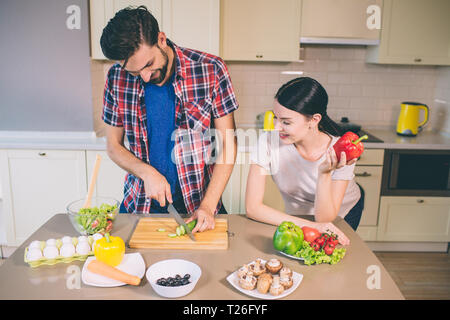 Happy couple is staying together. He cuts cucumber into pieces. She sits and looks at it. Girl holds red pepper in left hand. There are a lot of veget Stock Photo