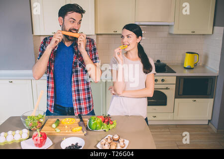 Hungry man stands and looks at girl. He devours carrot. Girl looks at vegetable. She bites a piece of yellow pepper. They look funny Stock Photo