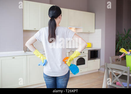 Busy woman stands in kitchen and looks at furniture. She wears yellow gloves. Girl has blue spray in one hand and rag in another one. She is ready to  Stock Photo