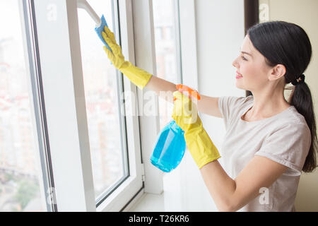 Nice and delightful girl stands in front of window and cleaning it with rag and blue liquid spray. Girl wears yellow gloves Stock Photo
