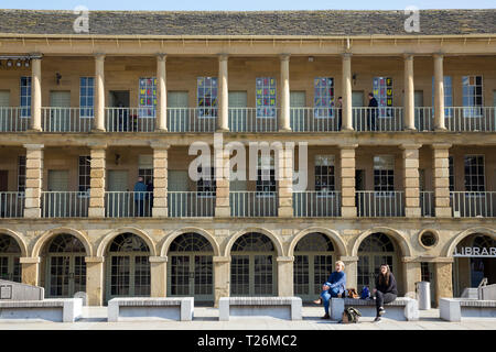 Women sitting on a bench in front of the east wall colonnade / colonnades / balcony / balconies of The Piece Hall. Sunny / sun & blue sky. Halifax, West Yorkshire, UK. (106) Stock Photo