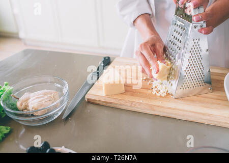 A picture of woman's hands grating piece of cheese on grater. It is on wood board. Knife is lying on table Stock Photo